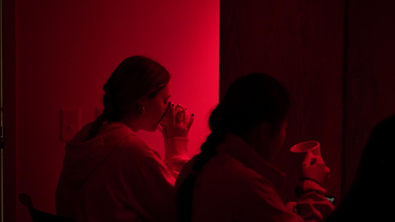 Two students sit in a room with dark red lighting as they sip from coffee cups