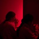 Two students sit in a room with dark red lighting as they sip from coffee cups
